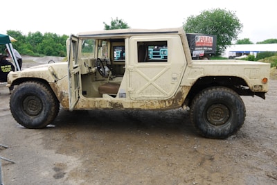 A rugged off-road vehicle with a beige exterior, covered in mud, is parked on a dirt path. The front door on the driver's side is open, revealing a simple interior with a steering wheel and seats. The vehicle has large, thick tires suitable for off-road terrain. In the background, there are some trees and a signboard, along with a cloudy sky.