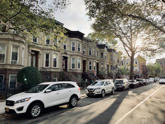 A row of historic brownstone buildings line a tree-shaded residential street. Sunlight filters through the leaves casting dappled shadows on the sidewalk and parked cars. Vehicles parked along the curb include white SUVs and other assorted cars. The overall scene conveys a peaceful and quiet neighborhood atmosphere.