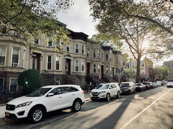 A row of historic brownstone buildings line a tree-shaded residential street. Sunlight filters through the leaves casting dappled shadows on the sidewalk and parked cars. Vehicles parked along the curb include white SUVs and other assorted cars. The overall scene conveys a peaceful and quiet neighborhood atmosphere.