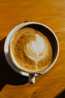 A cup of cappuccino with intricate latte art in the form of a leaf pattern is placed on a wooden surface. The foam is creamy and well-frothed, creating a contrast with the darker tones of the coffee beneath.