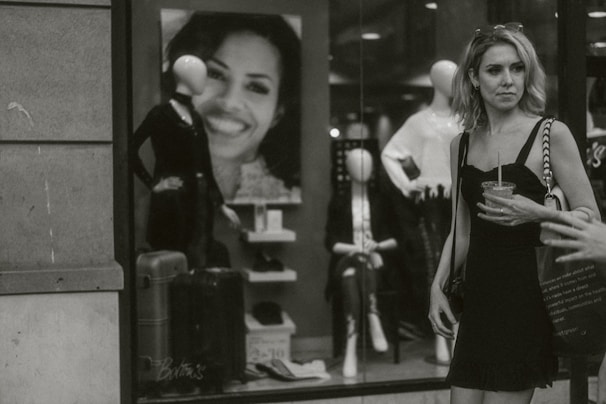 A chic woman in a classic black dress and pearl necklace, holding a coffee cup outside a stylish boutique.