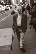 An elderly man with glasses is walking on a city sidewalk, using a cane. He is wearing a blazer, a polo shirt, and slacks. The background shows a busy street with blurred vehicles and pedestrians, creating a sense of urban life.