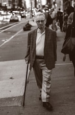 An elderly man with glasses is walking on a city sidewalk, using a cane. He is wearing a blazer, a polo shirt, and slacks. The background shows a busy street with blurred vehicles and pedestrians, creating a sense of urban life.