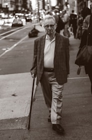 An elderly man with glasses is walking on a city sidewalk, using a cane. He is wearing a blazer, a polo shirt, and slacks. The background shows a busy street with blurred vehicles and pedestrians, creating a sense of urban life.