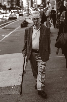 An elderly man with glasses is walking on a city sidewalk, using a cane. He is wearing a blazer, a polo shirt, and slacks. The background shows a busy street with blurred vehicles and pedestrians, creating a sense of urban life.