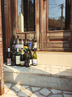 A collection of wine bottles is arranged on the steps in front of a wooden door. The door has a weathered appearance with peeling paint, suggesting age or rustic charm. Reflections can be seen in the glass panels of the door, and there is a mix of sunlight and shadow on the scene, indicating a clear day.