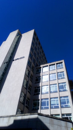 A tall, modern office building with multiple windows against a clear blue sky. The structure has clean lines and a minimalist design, with a sign that reads 'SPACES.' on the side. The building appears to be made of concrete or a similar material.