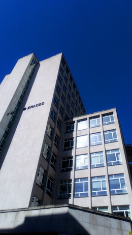 A tall, modern office building with multiple windows against a clear blue sky. The structure has clean lines and a minimalist design, with a sign that reads 'SPACES.' on the side. The building appears to be made of concrete or a similar material.