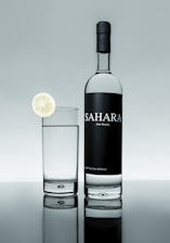 A bottle of Sahara date brandy is positioned next to a glass filled with a clear liquid. A slice of lime garnishes the rim of the glass. The background and surface create a reflective effect, giving a clean and sophisticated appearance.