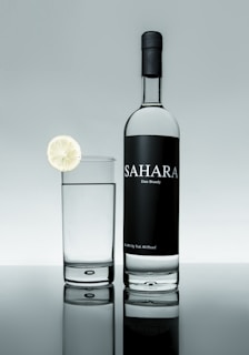 A bottle of Sahara date brandy is positioned next to a glass filled with a clear liquid. A slice of lime garnishes the rim of the glass. The background and surface create a reflective effect, giving a clean and sophisticated appearance.