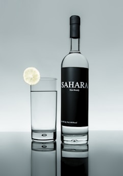 A bottle of Sahara date brandy is positioned next to a glass filled with a clear liquid. A slice of lime garnishes the rim of the glass. The background and surface create a reflective effect, giving a clean and sophisticated appearance.