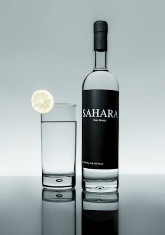 A bottle of Sahara date brandy is positioned next to a glass filled with a clear liquid. A slice of lime garnishes the rim of the glass. The background and surface create a reflective effect, giving a clean and sophisticated appearance.