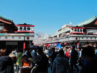 A bustling street scene features a crowd of people walking and shopping in a market area adorned with traditional Japanese signage and architecture. The sky is clear and blue, enhancing the vibrant atmosphere. Various stalls are set up selling goods under the green-roofed structures.