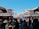 A bustling street scene features a crowd of people walking and shopping in a market area adorned with traditional Japanese signage and architecture. The sky is clear and blue, enhancing the vibrant atmosphere. Various stalls are set up selling goods under the green-roofed structures.