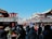 A bustling street scene features a crowd of people walking and shopping in a market area adorned with traditional Japanese signage and architecture. The sky is clear and blue, enhancing the vibrant atmosphere. Various stalls are set up selling goods under the green-roofed structures.