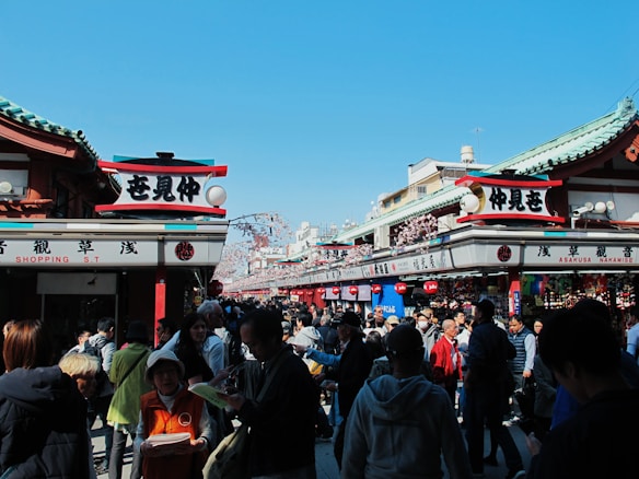 A bustling street scene features a crowd of people walking and shopping in a market area adorned with traditional Japanese signage and architecture. The sky is clear and blue, enhancing the vibrant atmosphere. Various stalls are set up selling goods under the green-roofed structures.