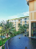 A modern luxury hotel with a contemporary architectural design features multiple stories with large glass windows and beige accents. Lush palm trees and well-maintained green gardens surround the building. The foreground showcases a tranquil infinity pool with a smooth, dark surface.