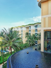 A modern luxury hotel with a contemporary architectural design features multiple stories with large glass windows and beige accents. Lush palm trees and well-maintained green gardens surround the building. The foreground showcases a tranquil infinity pool with a smooth, dark surface.