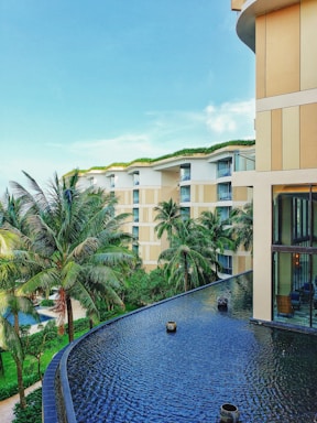 A modern luxury hotel with a contemporary architectural design features multiple stories with large glass windows and beige accents. Lush palm trees and well-maintained green gardens surround the building. The foreground showcases a tranquil infinity pool with a smooth, dark surface.