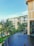 A modern luxury hotel with a contemporary architectural design features multiple stories with large glass windows and beige accents. Lush palm trees and well-maintained green gardens surround the building. The foreground showcases a tranquil infinity pool with a smooth, dark surface.