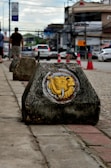 A painted concrete block is placed on a sidewalk with a yellow elephant design on it. The background shows a street scene with vehicles and pedestrians. Traffic cones are visible along the road, and there are buildings and overhead wires in the distance under a cloudy sky.