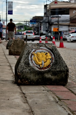 A painted concrete block is placed on a sidewalk with a yellow elephant design on it. The background shows a street scene with vehicles and pedestrians. Traffic cones are visible along the road, and there are buildings and overhead wires in the distance under a cloudy sky.