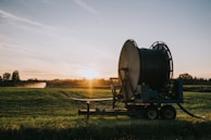 A large agricultural irrigation reel is positioned in a open field during sunset. The equipment is on a wheeled platform, and there are distant trees with an irrigation spray visible in the background.