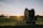 A large agricultural irrigation reel is positioned in a open field during sunset. The equipment is on a wheeled platform, and there are distant trees with an irrigation spray visible in the background.
