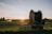 A large agricultural irrigation reel is positioned in a open field during sunset. The equipment is on a wheeled platform, and there are distant trees with an irrigation spray visible in the background.