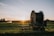 A large agricultural irrigation reel is positioned in a open field during sunset. The equipment is on a wheeled platform, and there are distant trees with an irrigation spray visible in the background.