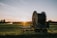A large agricultural irrigation reel is positioned in a open field during sunset. The equipment is on a wheeled platform, and there are distant trees with an irrigation spray visible in the background.