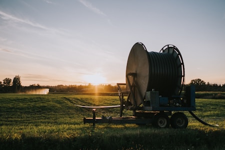 A large agricultural irrigation reel is positioned in a open field during sunset. The equipment is on a wheeled platform, and there are distant trees with an irrigation spray visible in the background.