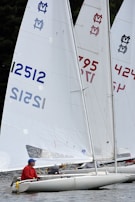 Several sailboats navigate close to each other on the water, each displaying prominent sails with unique numbers and logos. The scene involves individuals actively participating in a sailing activity, with one person in a red shirt and blue cap clearly visible.