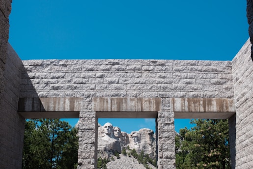 The image shows the Mount Rushmore sculpture framed by a stone archway. The sculptures of four U.S. presidents are carved into the mountain in the background, while the foreground features a textured stone structure. The clear blue sky contrasts with the gray stones and green trees surrounding the area.