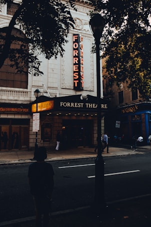 A historic theater with an ornate facade features the bright red vertical sign reading 'FORREST.' The marquee displays the sign for 'Hamilton.' A few people stand or walk on the street outside, with trees partially shading the view.