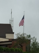 A building with a gabled roof, featuring an American flag prominently displayed on a flagpole. The surroundings include some trees and a cloudy sky. The structure has a combination of brick and light-colored walls, with a modern architectural design.