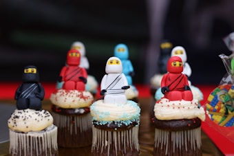Several cupcakes are topped with tiny figurines resembling ninjas in different colors, including black, red, blue, and white. Each figurine is sitting on a cupcake decorated with different kinds of sprinkles and frosting.
