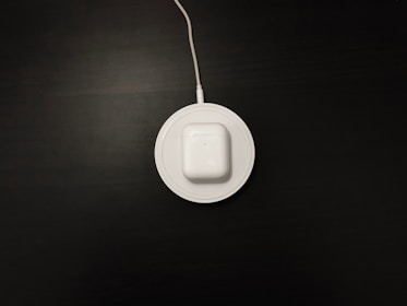 A white wireless charging pad with a pair of white earbuds in a case is situated on a dark surface. The charging pad is connected to a cable that extends out of the top of the image.