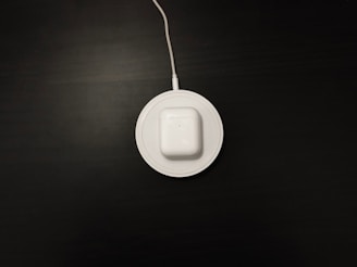 A white wireless charging pad with a pair of white earbuds in a case is situated on a dark surface. The charging pad is connected to a cable that extends out of the top of the image.