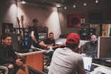 Inside a recording studio, several people are engaged in a relaxed and collaborative session. There are guitars positioned in a stand, a person with a baseball cap standing, two individuals sitting and conversing, one holding a guitar, and another working on a laptop. The room is equipped with audio equipment, including speakers and a mixing console.