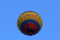 A colorful hot air balloon with a design featuring yellow, blue, and red stripes. The words 'reservoya.com' are visible on the side of the balloon. The background is a clear blue sky, emphasizing the brightness and vibrancy of the balloon's colors.