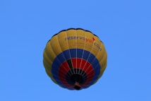 A colorful hot air balloon with a design featuring yellow, blue, and red stripes. The words 'reservoya.com' are visible on the side of the balloon. The background is a clear blue sky, emphasizing the brightness and vibrancy of the balloon's colors.