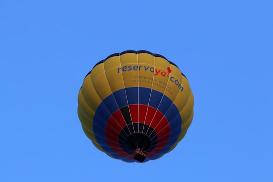 A colorful hot air balloon with a design featuring yellow, blue, and red stripes. The words 'reservoya.com' are visible on the side of the balloon. The background is a clear blue sky, emphasizing the brightness and vibrancy of the balloon's colors.