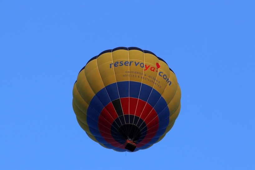 A colorful hot air balloon with a design featuring yellow, blue, and red stripes. The words 'reservoya.com' are visible on the side of the balloon. The background is a clear blue sky, emphasizing the brightness and vibrancy of the balloon's colors.
