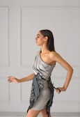 A woman with long dark hair is wearing a stylish, modern dress in soft gray tones. The dress features a unique asymmetrical design with intricate embroidered patterns in darker shades. She is posing gracefully against a minimalist light gray background with panel details.