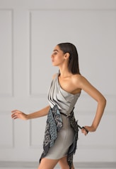 A woman with long dark hair is wearing a stylish, modern dress in soft gray tones. The dress features a unique asymmetrical design with intricate embroidered patterns in darker shades. She is posing gracefully against a minimalist light gray background with panel details.