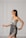 A woman with long dark hair is wearing a stylish, modern dress in soft gray tones. The dress features a unique asymmetrical design with intricate embroidered patterns in darker shades. She is posing gracefully against a minimalist light gray background with panel details.