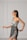 A woman with long dark hair is wearing a stylish, modern dress in soft gray tones. The dress features a unique asymmetrical design with intricate embroidered patterns in darker shades. She is posing gracefully against a minimalist light gray background with panel details.