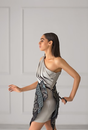 A woman with long dark hair is wearing a stylish, modern dress in soft gray tones. The dress features a unique asymmetrical design with intricate embroidered patterns in darker shades. She is posing gracefully against a minimalist light gray background with panel details.