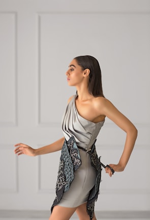 A woman with long dark hair is wearing a stylish, modern dress in soft gray tones. The dress features a unique asymmetrical design with intricate embroidered patterns in darker shades. She is posing gracefully against a minimalist light gray background with panel details.